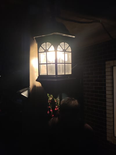 An outdoor lantern with a decorative arched window design emits a bright, warm light. The lantern is mounted on a wooden post. To the left of the lantern, a solar-powered security light with multiple LEDs is attached to the same post. In the foreground, a person's head and shoulders are visible, silhouetted against the light. To the right of the lantern, a brick wall with white trim is visible. A plant with red flowers hangs near the lantern. The overall scene suggests a nighttime setting, likely in a residential area in Alabino, Russia.