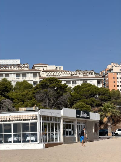A wide-angle, eye-level shot captures a beachfront scene in Urbanització Cales I Talaies, Spain, under a clear, bright blue sky. In the foreground, a sandy beach slopes gently towards a white, single-story building that houses a restaurant. The building features large glass windows, some of which offer a view of tables set with white tablecloths and blue chairs inside. An awning with a striped pattern and the letter "M" is visible above some of the windows. A man wearing a black t-shirt, blue shorts, and carrying a backpack walks away from the restaurant on the sand. To the right of the restaurant, a palm tree stands prominently. In the background, behind a dense line of green trees, are several multi-story buildings, including a large hotel with the word "VALLESOL" visible on its facade. Further to the right, a taller, brick-clad building rises against the sky. A black car is parked near the restaurant, and a white car is visible on a road behind it. Several small dark specks, possibly birds, are scattered across the sky.