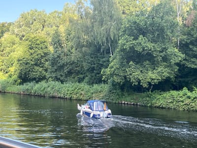 In Teltow, Germany, a small blue and white motorboat cruises down a canal, leaving a foamy wake on the dark, rippling water. The boat, with its enclosed cabin, appears to have at least two occupants. Along the canal's edge, lush green reeds and bushes form a dense barrier, transitioning into a backdrop of tall, mature trees that dominate the landscape. The sky above is a clear, pale blue. The sun casts a gentle light on the scene.