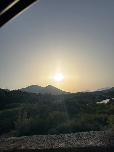 The image depicts a serene natural landscape under a bright, hazy sun, likely during sunrise or sunset. The sun, a radiant orb, is positioned in the upper center of the frame, casting a soft, diffused light across the scene. In the foreground, a rough, textured stone surface occupies the bottom edge, suggesting a viewpoint from a bridge or wall. Beyond this, rolling hills covered in lush, dark green vegetation dominate the mid-ground. A winding river or body of water is visible on the right side, reflecting the ambient light. Silhouettes of distant mountains are visible on the horizon, adding depth to the composition. A few scattered structures, appearing to be houses, are present on the left side, nestled amongst the trees, hinting at a nearby settlement. The overall atmosphere is tranquil and picturesque, characteristic of a Mediterranean environment, possibly near the city of Orosei, Italy, known for its scenic natural beauty.