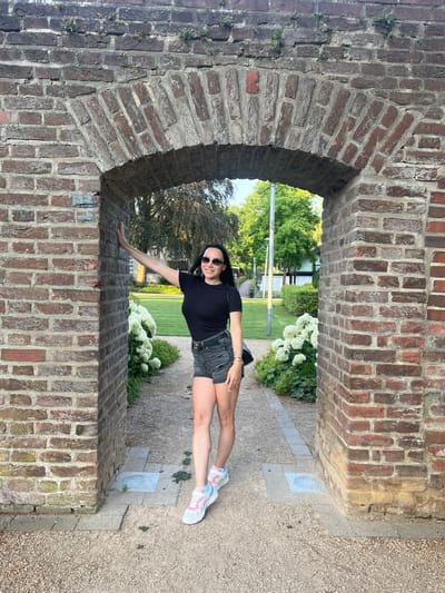 A full-body, eye-level, medium shot shows a young woman standing in a weathered brick archway. She is positioned in the center of the arch, facing forward, and smiling. Her right arm is extended and resting on the left pillar of the arch. She is wearing a black t-shirt, dark denim shorts, white sneakers with pink and blue accents, and sunglasses. A black purse is slung over her right shoulder. The archway is constructed from reddish-brown bricks and is partially covered in greenery. Beyond the arch, a pathway leads through a garden with white hydrangeas and lush green trees. The ground in the foreground and within the archway is covered in gravel and cobblestones. The scene appears to be in the gardens of Wassenberg Castle in Wassenberg, Germany.