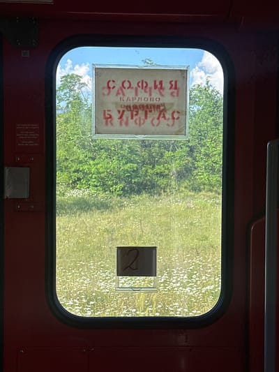 The image is an interior view from a train carriage window looking out at a field of wildflowers and trees under a blue sky with clouds. On the interior of the train door, there is a sign with Cyrillic text in red stencils. The text appears to read "СОФИЯ КАРЛОВО" on the top line and "БУРГАС" on the bottom line. These are likely destinations or routes. There are also smaller signs on the door with text in Bulgarian, English, and German stating "Do not open before the complete stop of the train" and "Nicht offnen bevor der Zug halt". A small sign with the number "2" is visible on the lower part of the window. The overall impression is that of a train carriage stopped or moving slowly through the Bulgarian countryside, near Karnare, a town known for its railway station.