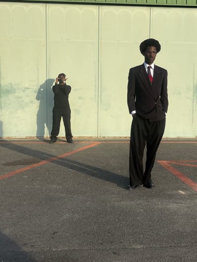 The scene depicts two individuals on an asphalt surface in Chonas-l'Amballan, France, against a pale green, corrugated metal wall. The ground features faded orange-red lines, including an 'X' pattern. Strong sunlight casts long shadows towards the viewer.
On the right, a Black male stands facing forward, wearing a dark double-breasted suit jacket over a white shirt and a red patterned tie, black wide-leg trousers, and dark shoes. He has an afro hairstyle and round eyeglasses, with his hands in his pockets.
On the left, a second Black male stands further back, holding a black smartphone to his eye, appearing to take a photograph or video. He wears a black long-sleeved top, black trousers, and dark shoes, with a gold-colored chain and a watch. His shadow is distinctly cast onto the pale green wall behind him.