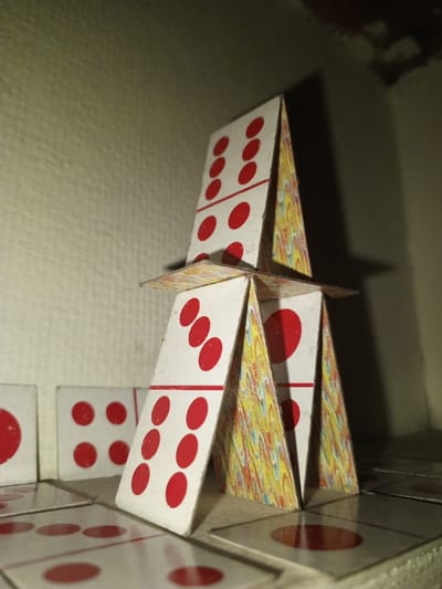 An assembled structure, resembling a house of cards, stands centrally on a flat, light-colored surface within an indoor setting in Kpein, Liberia. The structure is built from rectangular cards with white faces depicting red domino-style pips, separated by a red horizontal line, and backs featuring a yellow, red, and blue swirled pattern.
The base level consists of three cards forming an inverted 'V' shape, supporting a single horizontal card that acts as a platform. Two of these base cards are visible: one on the left displays a 3-pip and a 6-pip side, while the one on the right shows a single red pip on its visible side, its other half obscured. The patterned backs face inward.
Above this platform, two additional cards form a second inverted 'V'. The card on the left displays a 6-pip and a 6-pip side. The card on the right, also showing its patterned back inward, displays one visible 6-pip side.
Several other cards are scattered or arranged on the surface around the base of the structure. To the left, a card showing a single red pip lies flat, and another stands vertically, displaying four red pips on each half. Further back, another vertical card shows four red pips on each half. Partially visible cards with single pips and other configurations are on the foreground surface.
The background consists of a light beige or off-white wall with a subtle textured appearance, possibly painted or wallpapered. Illumination is uneven, casting shadows to the right and upper right of the structure, particularly from the upper card onto the wall. No individuals are present, and no active interactions are depicted.