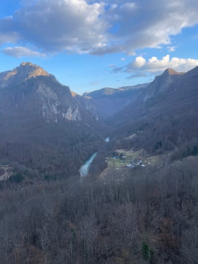 An aerial-perspective image captures a wide view of the Tara River Canyon in Đurđevića Tara, Montenegro, under a partly cloudy sky. The deep canyon, oriented generally from the upper center to the lower left, is dominated by steep, rugged mountainsides covered with bare deciduous trees, suggesting late autumn or winter. A turquoise-colored river, identified as the Tara River, snakes through the canyon floor. Sunlight illuminates the upper sections of the mountains, particularly a prominent peak on the left and ridges on the right, contrasting with the shadowed canyon depths. The sky is blue with scattered white and grey cumulus clouds. On the right bank of the river, in the mid-ground, a small settlement consisting of approximately seven structures with various roof colors, including orange and blue, is visible on a relatively flat area amidst the trees. The immediate foreground is comprised of the bare branches of trees, indicating a high vantage point. No individuals, animals, or specific actions are discernible.