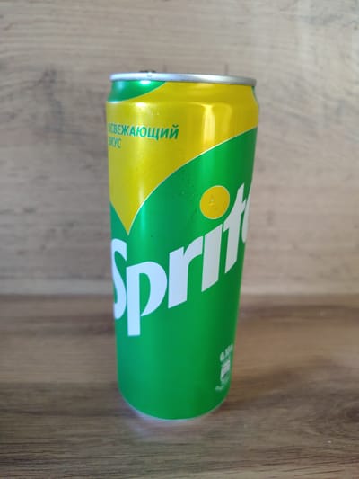 A close-up shot focuses on a green and yellow Sprite soda can, positioned vertically on a wooden surface. The can's label features prominent white "Sprite" lettering, with smaller text above reading "ОСВЕЖАЮЩИЙ ВКУС" (refreshing taste) in green. The background consists of a wooden wall, creating a simple, uncluttered setting. The lighting suggests an indoor environment. The scene is static and contains no people or overt actions. The image composition is centered on the can, emphasizing its branding and design.