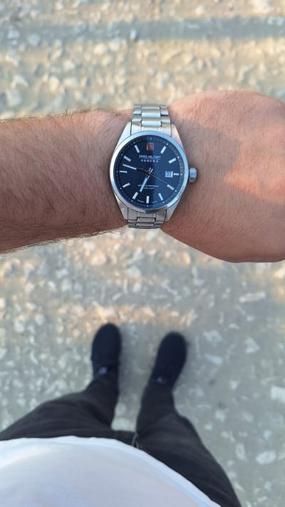 The image shows a close-up of a person's wrist wearing a Swiss Military Hanowa watch. The watch has a black face with silver indices and a date window displaying "30". The watch band is silver metal. The person's arm is hairy and is resting on a textured surface that appears to be gravel or small stones. In the background, the lower legs and feet of the person are visible, wearing black pants and black shoes. The ground is covered in the same gravelly texture. The image was taken in Sărata-Galbenă, Moldova.