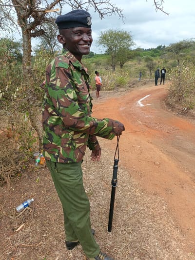 A smiling Kenyan soldier stands in a camo jacket, dark pants, and a black cap. He holds a black baton in his hand. A dirt road curves behind him with three people in the distance. A young person in a pink top and brown shorts is on the left of the road. Scattered trees and bushes dot the landscape under a cloudy sky. Litter is visible on the ground next to the soldier.