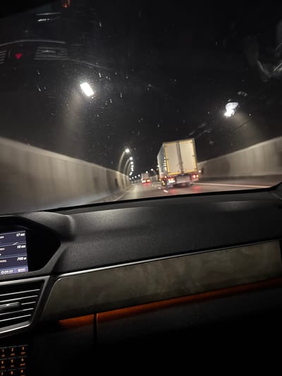 The image, taken from inside a vehicle, depicts a nighttime scene within a tunnel. The perspective is from the driver's seat, looking forward through the windshield. The tunnel is illuminated by artificial lights, creating streaks and reflections on the dirty windshield. Several trucks are visible ahead, with their brake lights glowing red. The tunnel walls are grey and appear to be made of concrete. On the right side of the image, part of the car's dashboard is visible, including a digital display showing the time as 09:35 and some text in Cyrillic script, likely indicating distances or navigation information. The text also includes "17 км", "10 км", and "700 м". The overall atmosphere is dark and enclosed, typical of driving through a long tunnel at night. The condition of the windshield suggests the car has been driven in potentially wet or dusty conditions. The specific location is identified as Dolna Vasilitsa, Bulgaria.