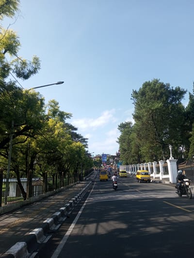 The image depicts a street scene in Nhot Chang, Laos. The road is asphalt with white-painted pedestrian crossings and yellow lines. Yellow vehicles, likely taxis, are moving along the road along with motorcycles. Sidewalks flank both sides of the road. Trees line both sides of the street, casting shadows on the road. A fence runs along the right side of the road. The sky is clear and blue. A streetlight is visible on the left. Billboards are visible in the distance. People are riding motorcycles.