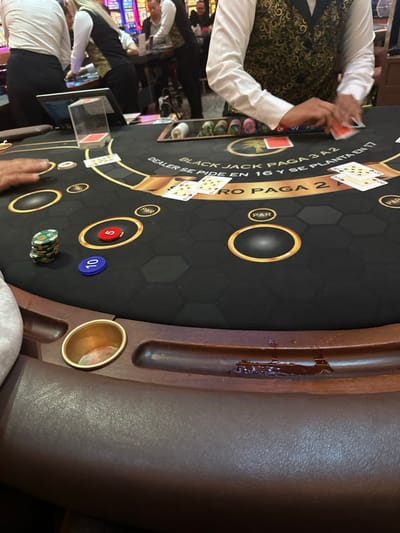 A blackjack table features a black surface with gold circular betting areas and hexagonal patterns. Two cards are dealt to a player, one face up and one face down. A dealer is in the process of distributing cards. Casino chips, including blue and red ones, are stacked on the table. A wooden railing surrounds the table's edge, with a cup holder built into it. Text on the table outlines the blackjack rules: "Black Jack pays 3 to 2," and "Dealer stands on 17."