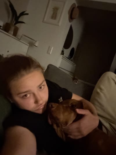 A person and a dog are lying down on a couch in a dimly lit room. The person is in the foreground, with their head and shoulders visible. They are looking at the camera with a neutral expression. The dog is in the person's arms, and its head is resting on the person's chest. The room appears to be a living room, with furniture and decorations visible in the background. The overall mood of the image is intimate and relaxed.