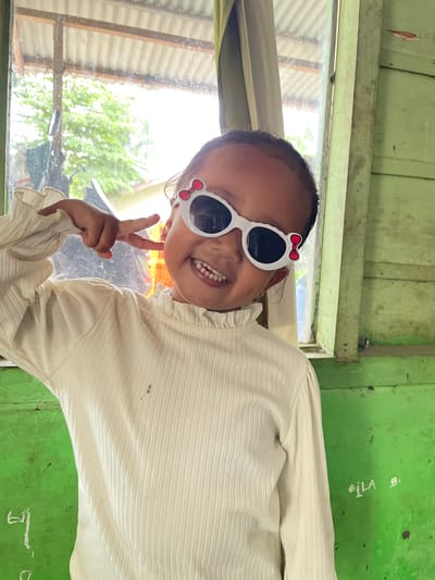 A young girl wearing white, long-sleeved shirt with a ruffled collar and white sunglasses adorned with red bows is posing for the camera indoors. She is smiling and making a peace sign with her right hand. The background shows a window with a view of trees and a rustic, green-painted wall with some writing on it. The setting appears to be a simple, possibly rural or underdeveloped area, consistent with the general appearance of Pantonrayeuk Satu, Indonesia.