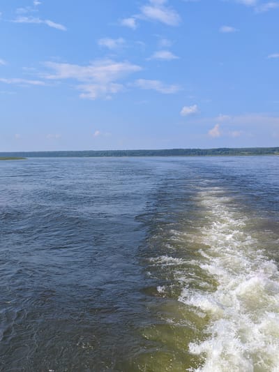 The image shows a wide, calm river with a wake from a boat in the foreground. The water is a dark blue-green color, with ripples and small waves. The wake is a frothy white, trailing behind the viewer. In the distance, a tree-lined shore is visible, with a hazy blue sky overhead dotted with white clouds. The scene appears to be a natural landscape, likely a river in a rural or undeveloped area. Without further information, it is not possible to specify the exact location in Potapovo, Russia.