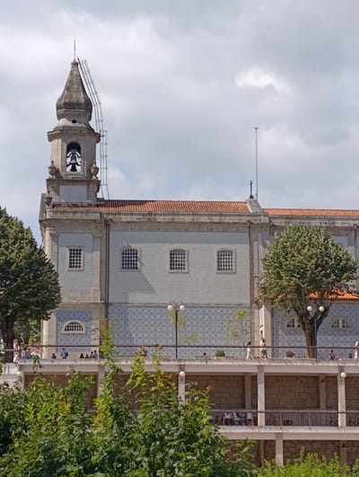 The image depicts the Santuário do São Bento das Peras, a sanctuary located in Terras de Bouro, Portugal. The sanctuary features a bell tower with a spire and a ladder attached to its side, suggesting ongoing maintenance or renovation. The main building has a tiled facade with a patterned design and a red-tiled roof. Several windows with intricate grilles are visible. A balcony or walkway runs along the front of the building, populated by a few individuals. Green trees are abundant in the foreground and on the sides of the building, framing the architectural structure. The sky is overcast with clouds.