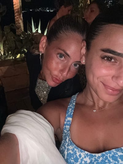 This nighttime outdoor image depicts two adult females posing for a photograph in Psarou, Greece. In the immediate foreground, positioned to the right, a female is visible from the chest up, showing her right profile and shoulder. She has dark hair pulled back, dark brown eyes, and a subtle closed-mouth smile. She wears a light blue and white patterned sleeveless top with thin straps and a delicate gold-colored necklace featuring a small circular pendant. A piece of white fabric partially covers her left shoulder. Behind and slightly to her left, a second female leans forward, looking directly towards the camera. She has lighter hair pulled back, light blue-green eyes, and a slight pouting expression. She wears a dark outer layer over a sparkling, possibly sequined, inner top. A long silver-colored dangle earring is visible on her left ear, along with a small stud nose piercing on her right nostril. Her left hand is raised, with fingers partially visible resting near her right temple, and a ring is present on her left ring finger. The background is dark, revealing distant city lights strung across what appears to be a body of water, consistent with a coastal view. Prominent spiky succulent plants, such as agave, are visible and artificially illuminated, casting strong silhouettes. A textured stone or pillar-like structure is partially visible on the far left. Blurry figures are discernible further in the dimly lit background, suggesting a social gathering.