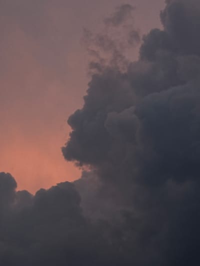 The image shows a dramatic sky filled with dark, ominous clouds against a soft, pinkish-orange sunset. The clouds are dense and voluminous, obscuring much of the sky. There is a stark contrast between the illuminated upper left portion of the sky and the shadowed, turbulent masses of clouds that dominate the right and lower parts of the frame. The texture of the clouds suggests movement and depth, with lighter areas hinting at reflected sunlight and darker areas indicating the thickness and density of the storm clouds. The overall mood is one of approaching weather and a sense of awe at the power of nature. The location is Savenki, Russia. There are no people, subjects, or objects visible in the image, only the sky.