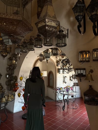 An indoor scene within a decorative lighting and artisan shop in Tlaquepaque, Mexico, is depicted. A single individual, seen from behind, stands in the center-left of the frame, facing into the shop's interior. This individual has dark, shoulder-length hair and is wearing a long-sleeved dark green dress with a pleated floor-length skirt, holding a clear bottle in their right hand.
The environment is characterized by a high density of metal lanterns and lamps of varying sizes and designs. Large, ornate, multi-faceted metal lanterns with intricate cutouts and glass panels hang from the ceiling. Numerous smaller lanterns are mounted on the light-colored, textured walls, functioning as sconces, some with visible price tags. Architectural features include several arched doorways and passageways leading into further sections of the establishment. The floor is composed of reddish-brown square tiles with light-colored grout lines.
In the middle-ground, a round glass-top table with an ornate black wrought-iron base displays multiple small, colorful decorative items, including what appear to be glass ornaments on wire trees. To the left, a smaller metal table holds a collection of brass bells. A dark, tree-like decorative stand adorned with yellow and orange heart-shaped glass ornaments is situated behind the individual. Illumination is provided by both natural light and the lit display lamps.