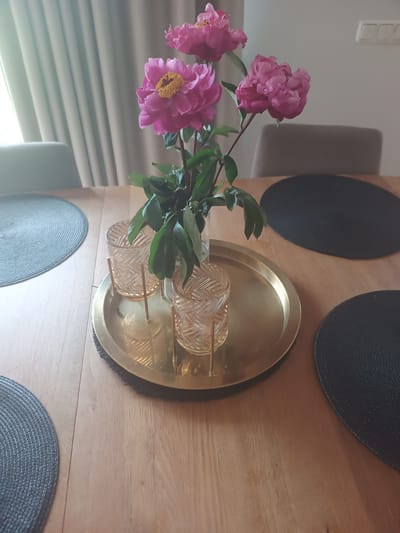 A close-up, eye-level shot shows a vase of pink peonies on a wooden dining table in Avižieniai, Lithuania. The vase, containing at least three pink peony blossoms with yellow centers and green leaves, sits on a round, gold-colored tray. Two decorative glass candle holders with a woven pattern and gold accents are also on the tray. The table is set with two black circular placemats and one round gray placemat. In the background, a sheer, light-colored curtain is visible, and the backs of two chairs are partially visible. The lighting suggests it is daytime.