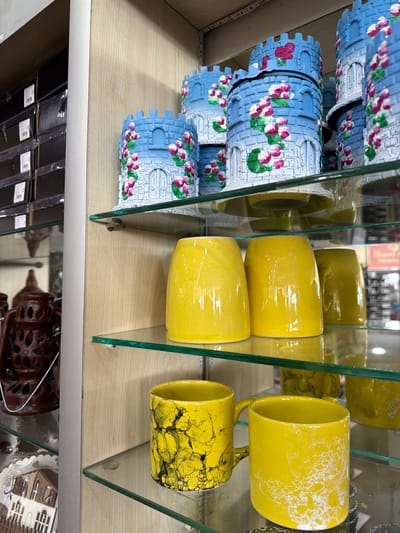 The image displays a retail display shelf in a store. The shelf is made of glass and contains various decorative items. On the top shelf, there are several cylindrical objects painted blue with white and pink floral designs, resembling castle towers. Below this, on the middle shelf, are two bright yellow ceramic cups or vases with a smooth, glossy finish. Further down, on the bottom shelf, two more yellow ceramic mugs are visible, one with a black and yellow marbled pattern, and another with a white speckled pattern on a yellow background. To the left of the shelves, a dark brown, intricately carved lantern-like object is partially visible, as well as a decorative plate depicting a building. The background shows a light-colored wooden paneling and some stacked boxes. The location is identified as Acıpayam, Turkey. There are no people or actions depicted in the image.