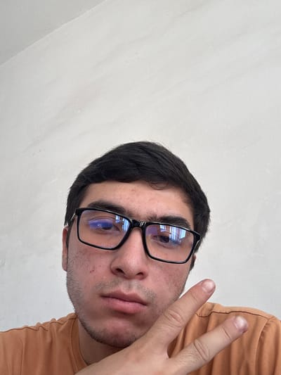 This is a close-up, selfie-style photograph of a young man wearing black-rimmed glasses and an orange t-shirt. He has dark hair and is looking towards the camera. His right hand is raised, with his index and middle fingers extended in a peace sign. The background is a plain, lightly textured off-white wall. The image appears to have been taken indoors. There is no visible information in the image indicating its location in Danghara, Uzbekistan.