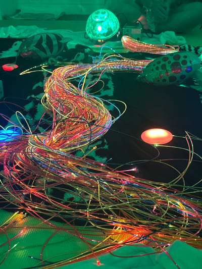 The image shows a close-up, high-angle view of a tangle of fiber optic cables illuminated with various colors, predominantly red, yellow, blue, and pink. The cables are spread across a dark surface, which appears to be a patterned fabric or blanket. In the background, several objects are visible, including a green disco ball, what appears to be a small electronic device, and two inflatable toys, one with black stripes and the other with pink spots. A red oval-shaped object is also illuminated in the foreground, possibly a light source. The overall impression is of a dimly lit, possibly playroom or sensory environment, with artificial lighting creating a colorful and somewhat chaotic visual effect. The location in Wakefield, United Kingdom, is not discernible from the image itself.