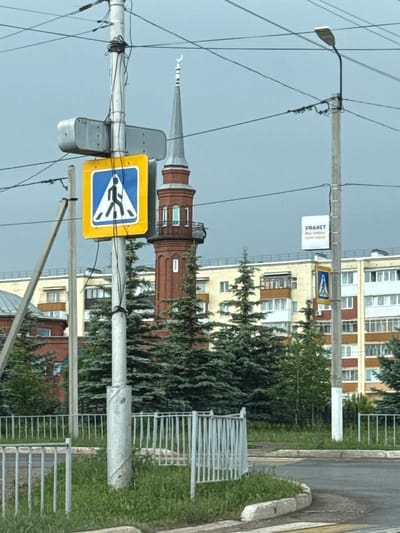 This image displays a daytime street scene in Belebei, Russia, under an overcast sky. In the foreground, an asphalt road featuring white pedestrian crossing stripes (zebra crossing) is bordered by concrete curbs and a low gray metal fence. A central gray utility pole, exhibiting a spiral wrapping around its base, supports numerous black electrical wires. Attached to this pole is a large yellow square sign with a blue triangular inset containing a black walking figure, indicating a pedestrian crossing. Above this sign, a gray rectangular utility enclosure is mounted. To the right, another gray utility pole features a curved streetlight arm and a white rectangular sign with red text that reads "УФАНЕТ Мы любим свой город!" (UFANET We love our city!).

In the midground, a red-brick minaret with a gray conical roof is visible, topped by a white crescent moon finial. The minaret features multiple rectangular windows and a balcony near its upper section. A dense cluster of dark green evergreen trees partially obscures the lower parts of the minaret and other structures. A smaller, identical pedestrian crossing sign is discernible further down the street to the right.

The background consists of several multi-story residential buildings with light-colored facades in shades of white, yellow, beige, and brown, equipped with numerous windows and balconies. The sky is a uniform light gray. No people are present in the image.