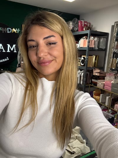 A woman with honey-blonde hair smiles in a selfie. She is wearing a white, ribbed top. Behind her, shelves display an assortment of packaged goods, possibly cosmetics or accessories. A circular sign with text, partially obscured, is visible on the lower left. The room seems to be an organized storage or workspace.