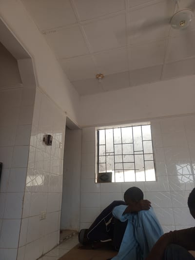 The image displays an interior room in Anguwar Jaba, Nigeria, with white tiled walls featuring a subtle geometric relief pattern and a white panelled ceiling. Two ceiling-mounted light fixtures are present: a central white ceiling fan with an integrated light, and a smaller, ornate gold and white lamp near the upper right corner.
A rectangular window, secured by a dark metal grid, is visible on the back wall, admitting significant natural light through its dirty panes. A small, dark, possibly decorative or religious item rests beneath the window. Electrical components are installed on the tiled wall to the left: a dark, rectangular control unit with a circular dial, and a white duplex electrical outlet below it. A smaller white switch or outlet is positioned to the right of the control unit. A decorative white molding runs horizontally along the top edge of the tiled walls.
The floor is covered by a light brown mat or rug. In the mid-ground, a young male individual with dark skin and short hair is seated on the mat, facing towards the left. He wears a light blue long-sleeved garment over dark trousers, and his right arm is bent, with his hand near his head, obscuring his face in a posture of rest or contemplation. Dark footwear, possibly sandals, is visible near his feet. Partially visible in the immediate foreground, on the right, is the upper back of another individual wearing a brown top and dark trousers. A doorway or archway to the left of the window reveals a darker area beyond.