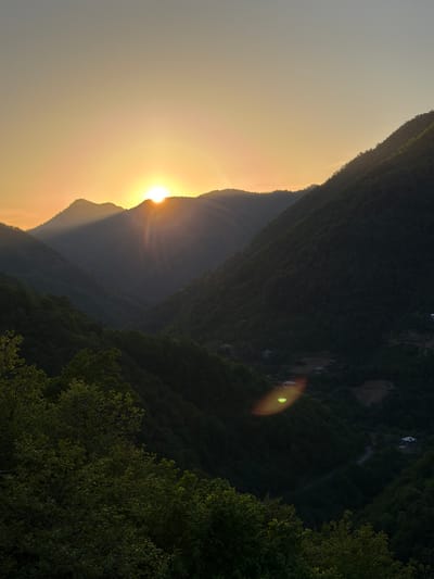 The image shows a mountainous landscape in what appears to be the city of Kveda Chkhutuneti, Georgia. The sun is setting behind a mountain, casting a golden glow across the sky and creating shadows on the mountain slopes. The scene includes dense trees, vegetation, and distant buildings, with a lens flare near the center of the image.