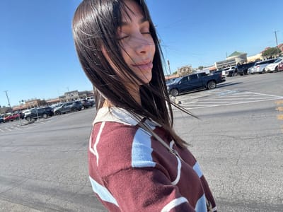 A woman with long, dark hair stands in a parking lot with her eyes closed, seemingly enjoying the sun. She wears a patterned burgundy and light blue sweater with a light-colored collar. The parking lot is filled with cars of various makes and models. A shopping center is visible in the background under a clear, blue sky.