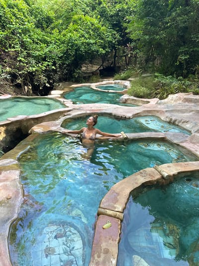 In Khlong Thom Nuea, Thailand, a woman with her hair in a bun is relaxing in a natural hot spring. She is wearing a leopard-print bikini and has her arms resting on the edge of the pool. The hot spring is composed of several tiered pools with rough, stone-like edges. The water is a clear, bright turquoise. The surrounding environment is lush with dense green foliage, creating a tropical atmosphere. In some of the pools, decorative tile patterns are visible beneath the water's surface, depicting stylized flora and fauna. The scene appears serene and natural.