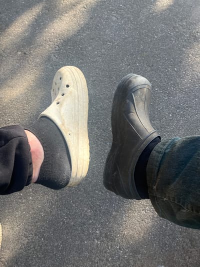 A close-up, overhead shot shows two feet on a paved surface, likely a sidewalk or road, with dappled sunlight casting shadows. The left foot wears a light-colored Croc with black socks and black pants. The right foot is clad in a dark-colored boot, also with black socks and dark denim pants. The boots and shoes appear to be dirty and well-worn, suggesting outdoor use. The environment is a neutral gray asphalt with small stones and debris visible. No specific landmarks in Серебрянский, Russia are identifiable.