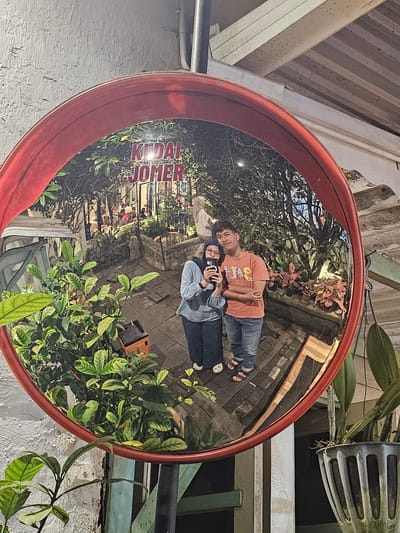 A convex mirror, mounted on a red frame, reflects a street scene in Parakan, Indonesia. The mirror, positioned outdoors, captures a glimpse of a "Kedai Jomer" establishment, with people seated at tables inside. A young couple is visible in the reflection, the woman holding a smartphone and taking a selfie with the man. The man wears an orange t-shirt and jeans, while the woman is in a blue top and hijab. Lush green foliage encroaches on the left side of the frame, partially obscuring the reflection. The background shows potted plants and stone landscaping, suggesting an outdoor seating area or garden. The overall lighting suggests it is evening.