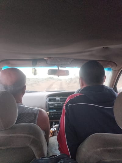 Two individuals are seated inside a vehicle. The person in the foreground, viewed from behind, is wearing a dark blue sweatshirt with red and white stripes on the sleeves and collar. They are seated in the driver's seat. To their left, the back of a bald man's head is visible. He is wearing a light-colored shirt. The vehicle's interior, including the dashboard with a car stereo and air vents, is visible. Through the windshield, a dusty, unpaved road stretches into the distance, suggesting a rural or desert environment. The sky is overcast. This scene is occurring in or near the city of Kokzhide, Kazakhstan.