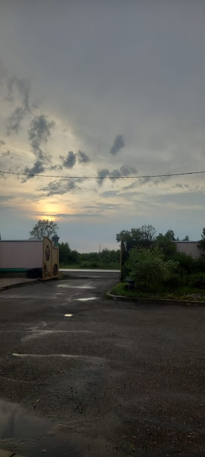 The image depicts a cloudy sky with the sun setting or rising, casting a warm glow through the clouds. The scene is outdoors, with a dark, wet asphalt road occupying the right side of the frame. Puddles of water reflect the sky and light on the road, indicating recent rain.

To the left of the road, there's a line of lush green trees and foliage. In the foreground, a building with a light-colored facade is visible, and to its left, a decorative metal structure, possibly a gate or part of an entrance, stands. The overall setting appears to be a somewhat rural or suburban area.

The time of day is clearly around sunrise or sunset due to the lighting and cloud formations. The weather seems to have recently been rainy or is still overcast with a chance of more rain. There are no people visible in the image. The mood is quiet and potentially serene, amplified by the soft light and the wetness of the ground. No text is visible in the image. The location context provided is Andzhero-Sudzhensk, Russia.