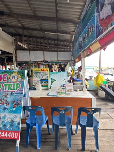 The image depicts a waterfront stall in Ban Phe, Thailand, likely a tourist information booth or souvenir shop. The stall is constructed of weathered wood and covered by a corrugated metal roof. Directly in front of the stall are three identical blue plastic chairs. Behind the chairs, a wooden counter displays various promotional materials, including posters advertising tourist activities like boat trips and jet skiing, as well as a map of local islands. One poster features the Thai text "มอเตอร์ไซค์ ให้เช่า" which translates to "Motorcycle for rent". Another poster is an "EXCHANGE MONEY" sign. A small Buddha statue sits on the counter, adorned with a yellow sash and flowers. A bright yellow plastic bag is prominent on the right side of the scene, along with a bicycle and various other objects suggesting a working commercial space. In the background, boats are moored in the water, with a distant bridge visible. The overall impression is one of a bustling, casual tourist hub.