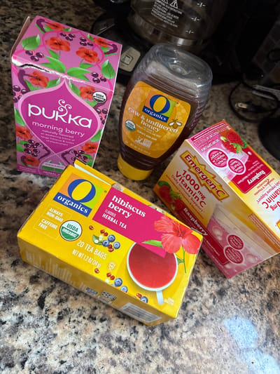 A high-angle shot captures a collection of health and wellness products on a countertop with a mottled beige and brown pattern, possibly granite. In the top left corner, the edge of a black coffee maker is visible with its power cord snaking down.
The items present, clockwise from the top, are: a box of Raspberry Emergen-C 1000mg Vitamin C packets; a box of "O Organics" Hibiscus Berry Herbal Tea containing 20 tea bags; an "O Organics" raw and unfiltered honey bottle; and a torn, partially crushed box of Pukka Morning Berry tea.
The setting appears to be a residential kitchen. There are no visible people or direct interactions.