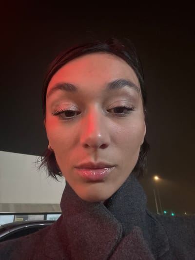 A person with light skin, dark hair, and glossy lips faces the camera at an angle. The lighting is dim, with a reddish hue on the nose and forehead, likely from an external light source. The individual is wearing a gray wool coat or jacket. The background appears to be a dark, possibly nighttime, setting. Hints of street lights and building elements are visible in the background. The person's expression is neutral.