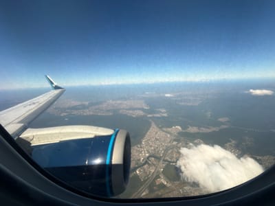 An aerial photograph, taken from the window of an airplane, captures a portion of the aircraft's exterior against a sprawling urban landscape under a clear sky. The left side of the frame is dominated by the aircraft's white wing, extending from the lower-left towards the upper-left corner. A winglet, featuring a light blue or teal tip, is visible at the end of the wing. Below the wing, a dark blue jet engine nacelle with a light grey front rim and a distinct light blue stripe is prominently featured in the bottom-left quadrant. Reflections are visible on the engine's surface. The dark, curved window frame partially borders the bottom of the image.
The sky, occupying the upper portion of the view, is a clear light blue with a gradual increase in atmospheric haze towards the horizon. Multiple white, cumulus clouds are present, with a significant cloud formation situated in the bottom-right, partially obscuring the ground below. The airplane window itself shows minor imperfections, such as small specks and a fine textured pattern, particularly visible against the sky.
Below the clouds and haze, an extensive and densely developed urban and suburban environment unfolds. This landscape features numerous buildings, an intricate network of roads and highways, and several bridges spanning various water bodies. A prominent, winding river or canal system is clearly visible, dissecting the urban area in the central-right. Interspersed with the development are patches of green vegetation, with larger forested areas appearing in the distant background, particularly towards the upper-right. Based on the distinctive estuarine geography, dense urban development, and major port infrastructure, the location is identified as the city of Santos, Brazil, specifically showing areas around the Port of Santos and São Vicente Island. The aircraft's livery on the engine and winglet are consistent with an Embraer E-Jet, commonly operated by Azul Linhas Aéreas. No people are visible. The scene is illuminated by natural daylight.
