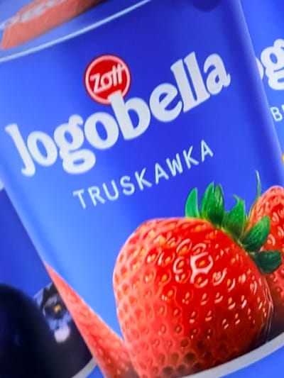 The image is a close-up, angled shot of a blue yogurt container, likely from a product called "Jogobella." The container features a strawberry and the word "TRUSKAWKA," which is Polish for strawberry. A circular red logo with "Zott" in white text is visible. The lighting is bright, suggesting it could be daytime. The background is a solid blue, with hints of other product packaging visible to the left and right, indicating a retail or promotional setting. There are no people or animals in the image. The focus is on the product packaging and the strawberry imagery.