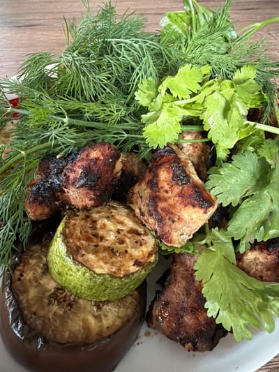 This media file is a close-up, top-down photograph of a plate of grilled food, likely a meal from Shashmurka, Russia. The primary subjects are pieces of grilled meat, possibly shashlik or kebab, alongside grilled slices of zucchini and eggplant. The meat appears to be seasoned and has char marks from grilling. The vegetables are also visibly grilled, with the zucchini having a slightly browned exterior and visible seeds, and the eggplant appearing soft and cooked through.

Surrounding the grilled items is a generous garnish of fresh green herbs. These include feathery dill and broad-leafed parsley or cilantro, adding a vibrant contrast to the cooked food. The herbs are fresh and appear to be slightly moist, suggesting they were recently washed.

The food is presented on a white plate, which is partially visible. The background shows a wooden surface, likely a table, adding a rustic element to the scene. There is no visible text in the image. The lighting suggests an indoor setting, possibly a kitchen or dining area, with soft illumination that highlights the textures and colors of the food. The overall impression is of a delicious and freshly prepared meal, typical of home-style cooking. The image does not depict any people or indicate a specific time of day or weather conditions beyond the general setting.
