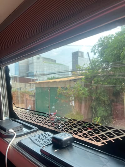 From the perspective of the interior of a vehicle, the image shows a view through a window, possibly a sunroof or side window, overlooking a street scene in Thôn 8, Vietnam. The foreground features part of the car's interior, including a brown shade partially covering the window, a black ventilation panel with triangular cutouts, and electronic devices connected with a white cable. The window reveals an exterior scene with a mix of greenery from trees, a building with a white façade, and structures with green and brown exteriors. Wires crisscross the sky above the buildings, suggesting an urban or suburban environment. There are no people clearly visible in the frame, but the presence of buildings and infrastructure indicates human habitation.
