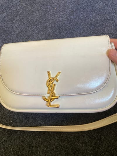A white, rectangular handbag with a gold interlocking "YSL" logo is centered in the frame. The handbag has a curved flap closure and a thin, light-colored strap. The material appears to be leather and shows some minor scuff marks and signs of wear. The handbag rests on a dark gray, textured surface, possibly a carpet or rug. The background is out of focus, but the surface appears consistent with the foreground. There is a human hand visible on the right side of the image, lightly touching the edge of the handbag. No other people or significant environmental details are visible. The location in Arbagish, Uzbekistan is not discernible from the image.