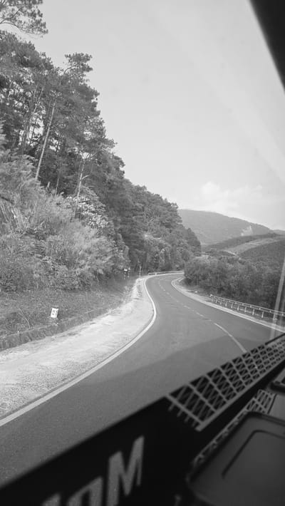 A black and white, slightly tilted image shows a winding road cutting through a lush, mountainous landscape. The road, paved and marked with a white line, curves sharply, hinting at steep inclines and declines. Dense vegetation blankets the mountain slopes, transitioning from tall trees to thick shrubbery closer to the road. Metal guardrails run alongside sections of the road, providing safety barriers. A road sign is partially visible near the roadside edge. The location is presumed to be Thôn 18, Vietnam, based on the prompt.
