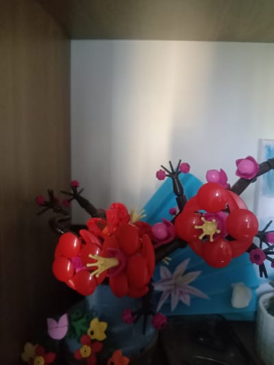 This image shows a close-up of decorative plastic flowers, likely part of a craft project or toy. The flowers are primarily red and pink, with yellow accents in the center of some. They are arranged on dark brown stems, mimicking branches. In the background, a light blue object with a white floral design is visible, along with a white wall and a dark brown shelving unit. There is no indication of location within the city of Kielce, Poland.