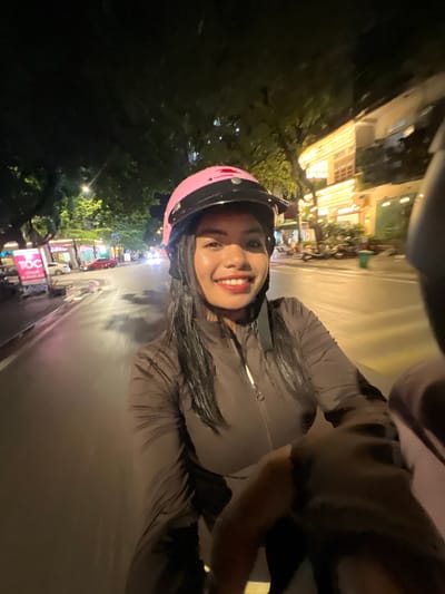 A woman wearing a pink helmet and a dark jacket rides a motorbike at night in Minh Xuân Ward, Vietnam. She is looking at the camera and smiling. The street is wet, and there are lights from buildings and streetlights. There are other vehicles on the road, including a red car. The woman's hair is wet, suggesting it may have rained recently or she has recently come from a wet environment.