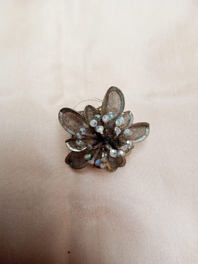 A single metallic brooch, designed in a floral motif, is depicted resting on a light, subtly textured fabric surface. The brooch consists of multiple layered petals fabricated from dark, fine-gauge metal mesh, exhibiting signs of tarnish or discoloration. Adorning the central area and interspersed among the petals are numerous small, faceted, spherical beads, primarily clear or translucent, which display an iridescent effect with discernible flashes of blue, green, and pink. Several dark, fibrous strands are present on the upper section of the brooch. The background is a uniform pale beige or cream-colored fabric, showing minor surface undulations and creases. No human figures, actions, or interactions are visible. The documented location is Qarabucaq qəsəbə, Azerbaijan.