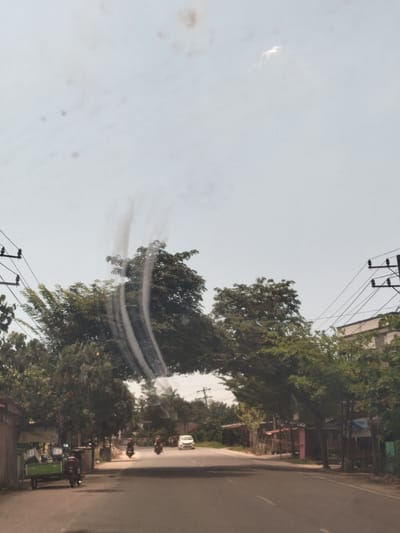 The image presents a street scene in Panteepanah, Indonesia, viewed from inside a vehicle with a slightly obscured windshield. A paved road stretches into the distance, flanked by trees and buildings on both sides. Power lines are visible overhead. There is a green stall and parked motorcycle on the left side of the road. Two motorbikes are moving down the road toward the camera. A white car is further down the road.