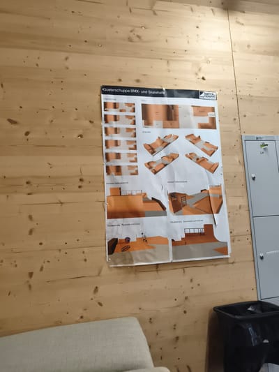 The image shows a poster on a wooden wall, likely in a skate hall or a similar facility in Wädenswil, Switzerland. The poster displays design concepts and renderings for a BMX and skate hall, with sections labeled "Arbeitsabschnitte," "Vision," "Ansichten," "Vorderer Hallenabschnitt," "Hinterer Hallenabschnitt," and "Visualisierung Pyramide und Curvy." The renderings depict various ramps, quarterpipes, and other skate park features. To the right of the poster, a gray locker bank is visible, with one locker door marked with "SK" in black marker. In the lower right corner, a black trash bin with a plastic liner is present. A portion of a light-colored sofa or chair is visible in the lower left corner. The overall environment appears to be an indoor space with a wooden planked wall.